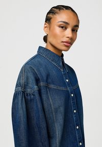 Chemise en denim bleu foncé avec un devant boutonné, un col structuré et des manches bouffantes, avec des coutures contrastées et une texture douce.