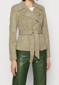 Veste de moto en simili cuir beige clair avec une taille ceinturée, des poches zippées et des détails en relief sur les manches.