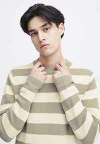 Beige og oliven stripet strikkegenser med rund hals og lange ermer. Teksturet stoff med kontrasterende brede horisontale striper.