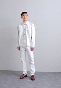 Veste en denim blanc avec des fermetures à bouton et des poches poitrine, assortie à un pantalon blanc. Des bottines en cuir marron viennent compléter la tenue.