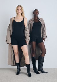 Two models wear black bodysuits; one in a long beige trench coat. Both have black knee-high boots. The setting is minimalist with light gray walls.