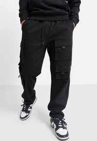 Persona vestida con pantalones cargo negros con múltiples bolsillos con solapa y zapatillas blancas y negras, con las manos en los bolsillos, frente a un fondo liso.