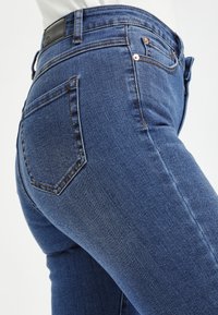 Jeans en denim bleu foncé avec une coupe ajustée. Les caractéristiques comprennent des poches arrière, une étiquette en cuir à la taille et des accents de couture visibles.