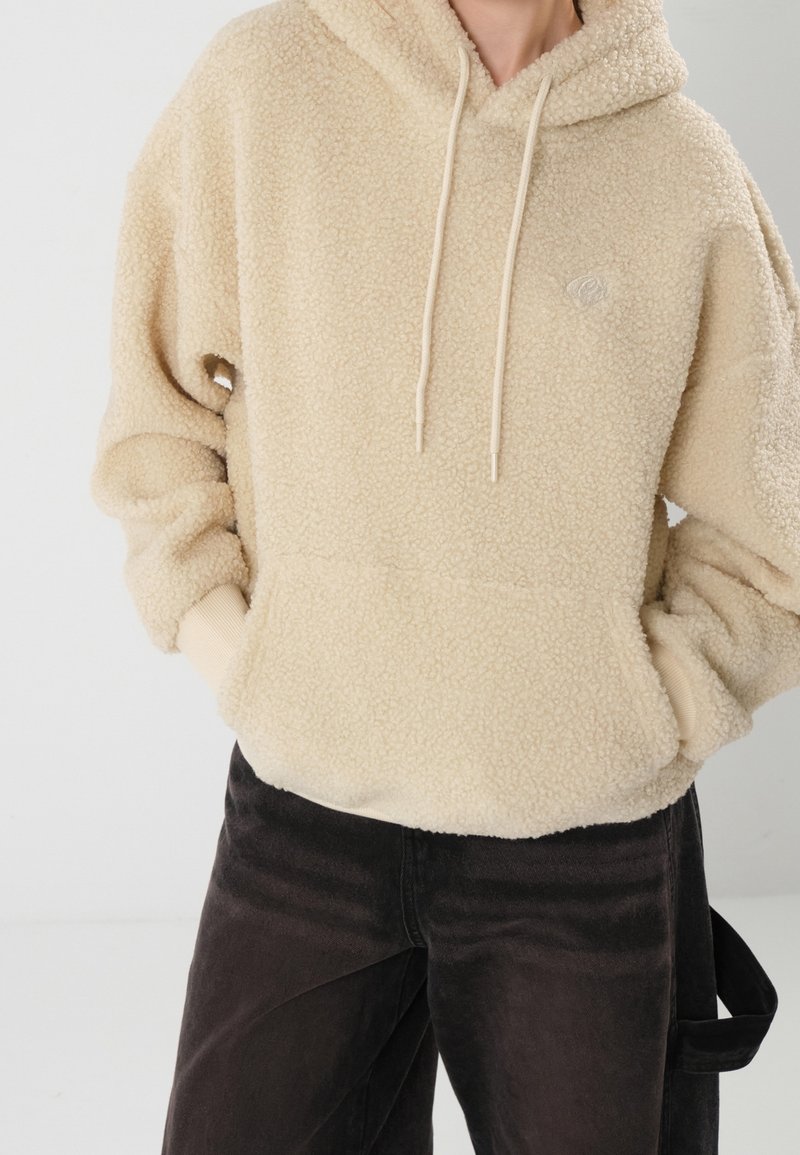 Personne portant un sweat à capuche en polaire beige texturée avec des cordons et un pantalon noir, les mains dans la poche avant du sweat.