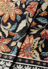 Černá látka se složitými květinovými vzory v oranžové, červené, žluté a modré barvě. Obsahuje dekorativní okraj s kulatými akcenty.