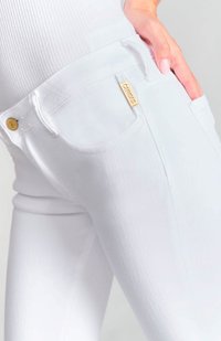 Jeans blancos con una textura suave, que presentan un bolsillo frontal y un acento de logo en tono dorado en la cinturilla. Costuras de mezclilla sutilmente visibles.