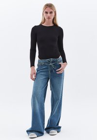 Schwarzer Langarm-Bodysuit kombiniert mit weit geschnittenen blauen Jeans, die einen gebundenen Bund, leichtes Ausbleichen und eine lockere Passform aufweisen. Weiße Sneakers sind inklusive.