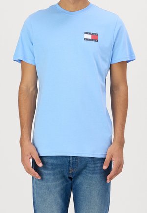 Mężczyzna noszący jasnoniebieską koszulkę Tommy Jeans z małym logo na piersi oraz niebieskie dżinsy, stojący na białym tle.