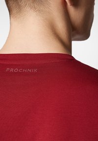 Czerwona koszula z krótkim rękawem o gładkiej fakturze i subtelnym prążkowaniu. Na plecach, blisko dekoltu, znajduje się logo "PROCHNIK".