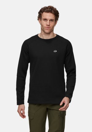 Man wearing black textured long-sleeve shirt with small embroidered logo on chest and olive green cargo pants against white background.