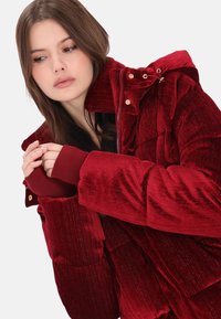 Piros steppelt dzseki, texturált, bordázott mintázattal, kapucnival, cipzáras lezárással és elasztikus mandzsettákkal. Aranyszínű fémrészletek láthatók.