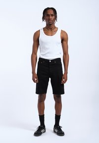 Shorts de mezclilla negra con un diseño ajustado, bolsillos frontales y cierre de botón, combinados con una camiseta sin mangas blanca y zapatillas negras.