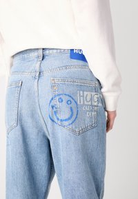 Light blue denim jeans with a relaxed fit. Features back pockets with a blue smiley face graphic and "HUG" text. Tag in blue fabric.