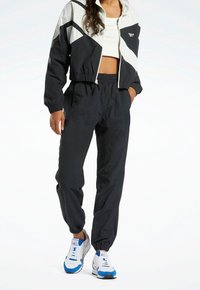 Svart och grå träningsdräkt med en kort jacka och byxor med elastisk midja. Har en geometrisk design, lätt material och sneakers med blå detaljer.