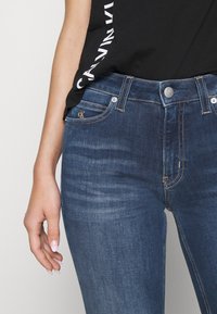 Vaqueros ajustados azul oscuro con un ligero desvanecimiento, que cuentan con una bragueta de botones y cremallera, y un logo de CK en el bolsillo delantero. Textura de denim suave.