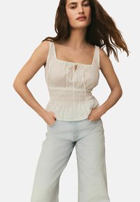 Haut sans manches blanc avec décolleté carrée, bodice froncé et détail à nouer, associé à un jean ample bleu clair. Tissu doux, texture subtile.