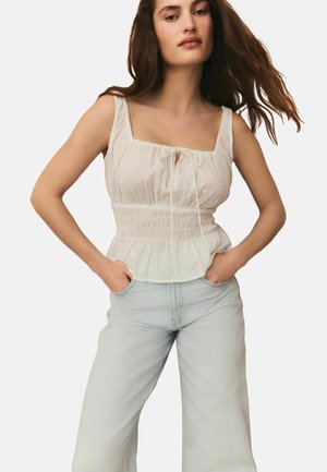 Witte mouwloze top met vierkante halslijn, geplooide lijf en strikdetail, gecombineerd met lichtblauwe wijde jeans. Zachte stof, subtiele textuur.