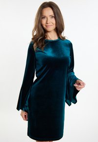Tengerzöld bársonydressz hosszú, bővülő ujjakkal és kerek nyakkivágással. Sima textúrával és térd alá érő fazonnal rendelkezik, laza illeszkedéssel.