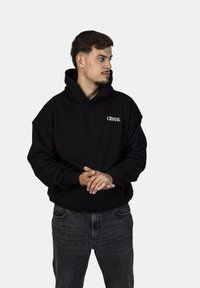 Felpa nera con cappuccio e testo "CRYSTAL" sul petto, design bulky, texture morbida, abbinata a jeans scuri. Il modello si trova in una posa neutra.