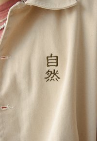 Beige bomullsskjorta med krage, med grön broderad text på bröstet. Tyget har ett slätt, strukturerat mönster.