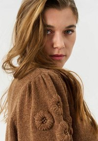 Brauner gestrickter Pullover mit floralen Stickereien an den Ärmeln, der ein strukturiertes Muster und weichen, warmen Stoff aufweist. Langes Haar fällt sanft.