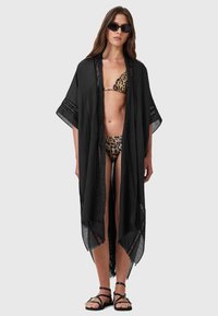 Svart, lätt kaftan med öppen front, lösa ärmar och spetsdetaljer, bärs över en leopardmönstrad bikini. Remsandaler fulländar looken.