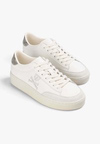 Zapatillas blancas de caña baja con suelas crema, cordones blancos, puntas de ante gris, detalles plateados en el talón y bordado de calaveras y tibias.