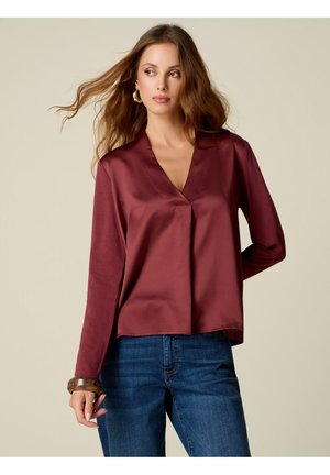 Frau mit langen Haaren, die ein burgunderfarbenes Satin-Oberteil mit langen Ärmeln, blaue Jeans, goldene Creolen und ein braunes Armband vor einfachem Hintergrund trägt.