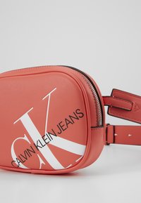Borsa a tracolla in pelle sintetica rossa con chiusura a zip, caratterizzata da un grande logo bianco "CK" e dalla scritta "CALVIN KLEIN JEANS" sul davanti.
