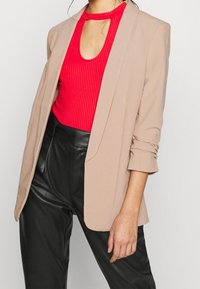 Blazer beige avec une coupe décontractée, doté d'un col à revers et de manches retroussées. Porté sur un top rouge à côtes et un pantalon en cuir noir.