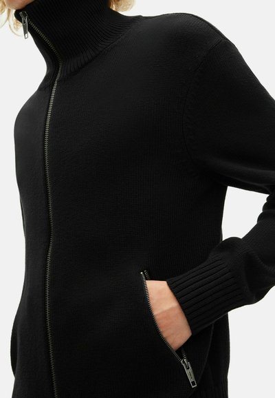 Personne portant un pull zippé noir avec des poignets côtelés, un col montant et une main dans une poche zippée sur le côté.