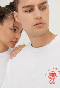 Hvid bomulds T-shirt med et rødt grafisk print med en globe karakter og tekst der siger "MOVING TO THE B." Korte ærmer, rund hals.