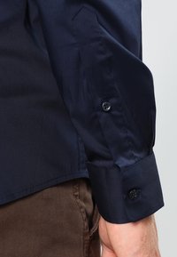 Navy blue long-sleeve shirt featuring a smooth texture, button cuffs, and structured shoulder seams. Fabric appears lightweight and wrinkle-resistant.
