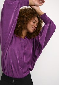 Blusa di raso viola con maniche a sbuffo e dettagli a bottoni. Il tessuto ha una trama liscia, in contrasto con una fascia in vita nera.