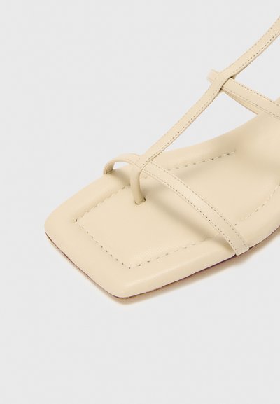 Sandalia de piel beige con punta cuadrada, que presenta finas correas cruzadas y costuras visibles a lo largo de los bordes. Textura suave y diseño minimalista.