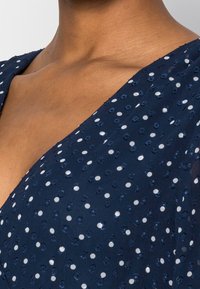 Laivansinis mekko, jossa on v-pääntie, pistekuvio valkoisilla ja hopeisilla yksityiskohdilla, teksturoitu kangas ja muotoiltu bodice-muotoilu.