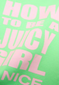 Un tessuto verde brillante presenta un grande testo rosa con la scritta "COME ESSERE UNA RAGAZZA JUICY NICE". Il materiale ha una texture a coste.