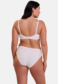Femme aux cheveux longs et foncés portant un soutien-gorge rose pâle et des culottes taille haute assorties, vue de dos contre un fond neutre.