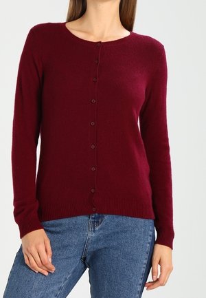 Frau trägt einen geknöpften, langärmligen, burgunderroten Cardigan und blaue Jeans, steht vor einem schlichten, hellen Hintergrund.