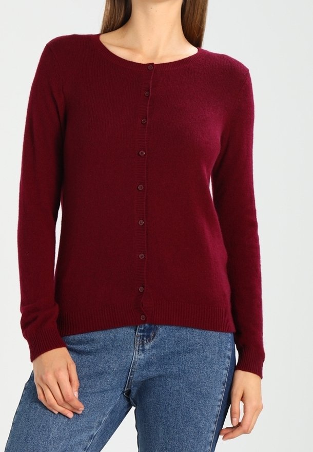 Femme portant un cardigan bordeaux boutonné à manches longues et un jean bleu, debout devant un fond clair uni.