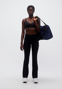 Γυναίκα με μαύρο αθλητικό μπουστάκι και παντελόνι κρατάει σκούρο μπλε τσάντα γυμναστηρίου στον ώμο, στέκεται μπροστά σε απλό λευκό φόντο.