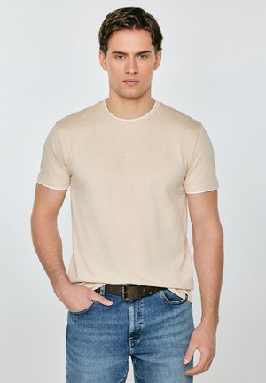 Noor mees heleda beež t-särgiga, mis on pandud siniste teksade sisse, seisab ühe käega taskus tavalise tausta ees.