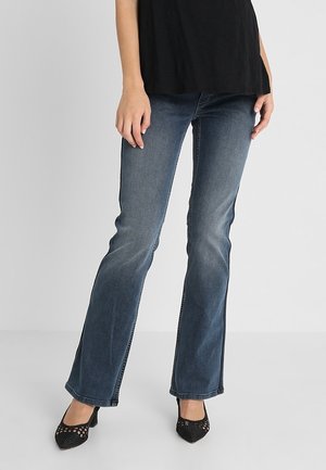 Vrouw draagt versleten blauwe bootcut jeans, zwart topje en zwarte pumps met spitse neus en mesh, staand op een lichtgrijze vloer.