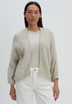 Woman wearing light beige open-front knit cardigan, ribbed beige top, and high-waist white pants against plain background.
