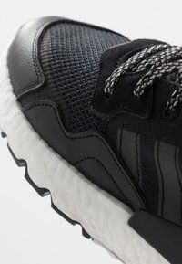 Svart sportssko med vit texturerad sula, mesh-ovandel, överdrag av mocka och svart-vita mönstrade snören på en neutral bakgrund.