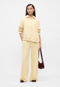 Ανοιχτό κίτρινο φούτερ με γιακά και κουμπιά, σε συνδυασμό με παντελόνι σε φαρδιά γραμμή. Κρατάει μια μικρή σκούρα μπορντό τσάντα.