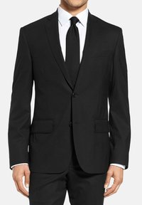 Veste noire avec deux boutons, revers crantés et poches latérales. Cravate texturée et chemise blanche avec col visible. Ajustement sur mesure.
