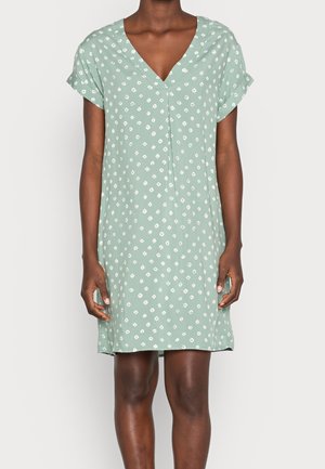 Vrouw met een lichtgroene jurk met korte mouwen en witte diamantvormige patronen, staande tegen een effen achtergrond.