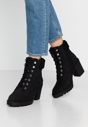 Bottines noires à lacets avec talons blocs et garniture en fourrure portées avec un jean bleu coupé au-dessus de la cheville sur un sol blanc.