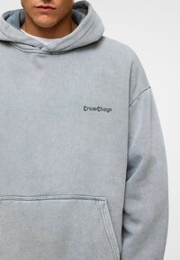 Lichtgrijze hoodie met een kangoeroezak, voorzien van de tekst "CreateChange" in cursief aan de linkerkant. Zacht, gestructureerd materiaal.